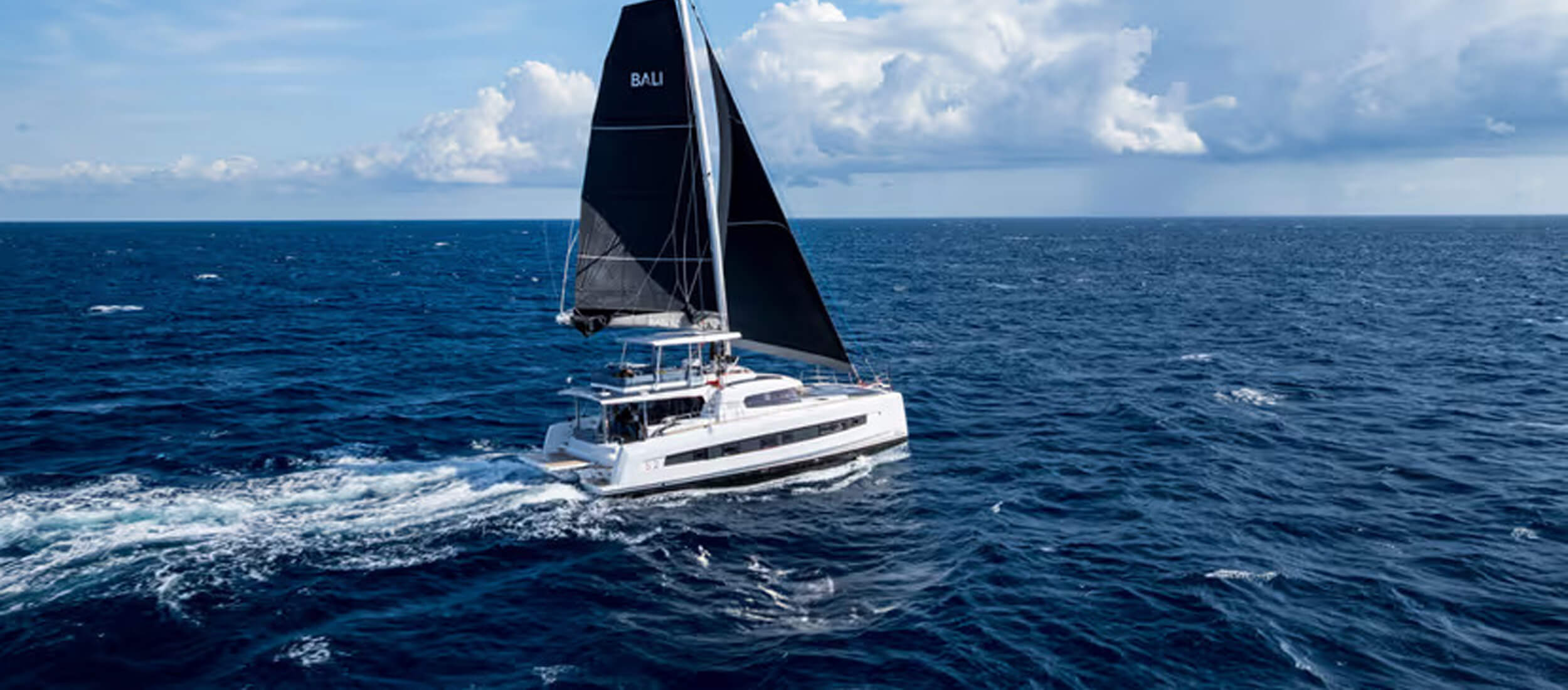 Athenian Yachts-Aerial view of a white sailing catamaran with dark sails cruising on the open sea under a cloudy sky.-Bali Catamarans for Sale