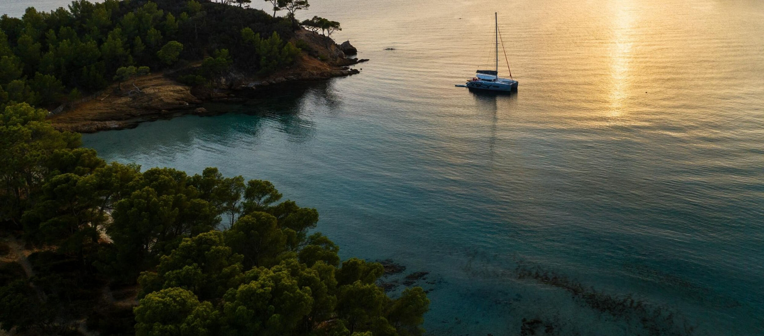 Athenian Yachts-A catamaran anchored in a calm bay near a wooded coastline, with sunlight shimmering across the surface of the water at sunset.-Excess Catamarans for Sale