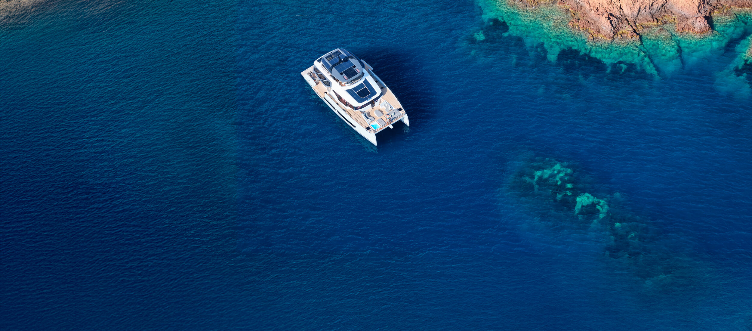 Athenian Yachts-Aerial view of a white catamaran anchored in deep blue open water near a rocky coastline with clear turquoise shallows visible below.-Fountaine Pajot Catamarans for Sale