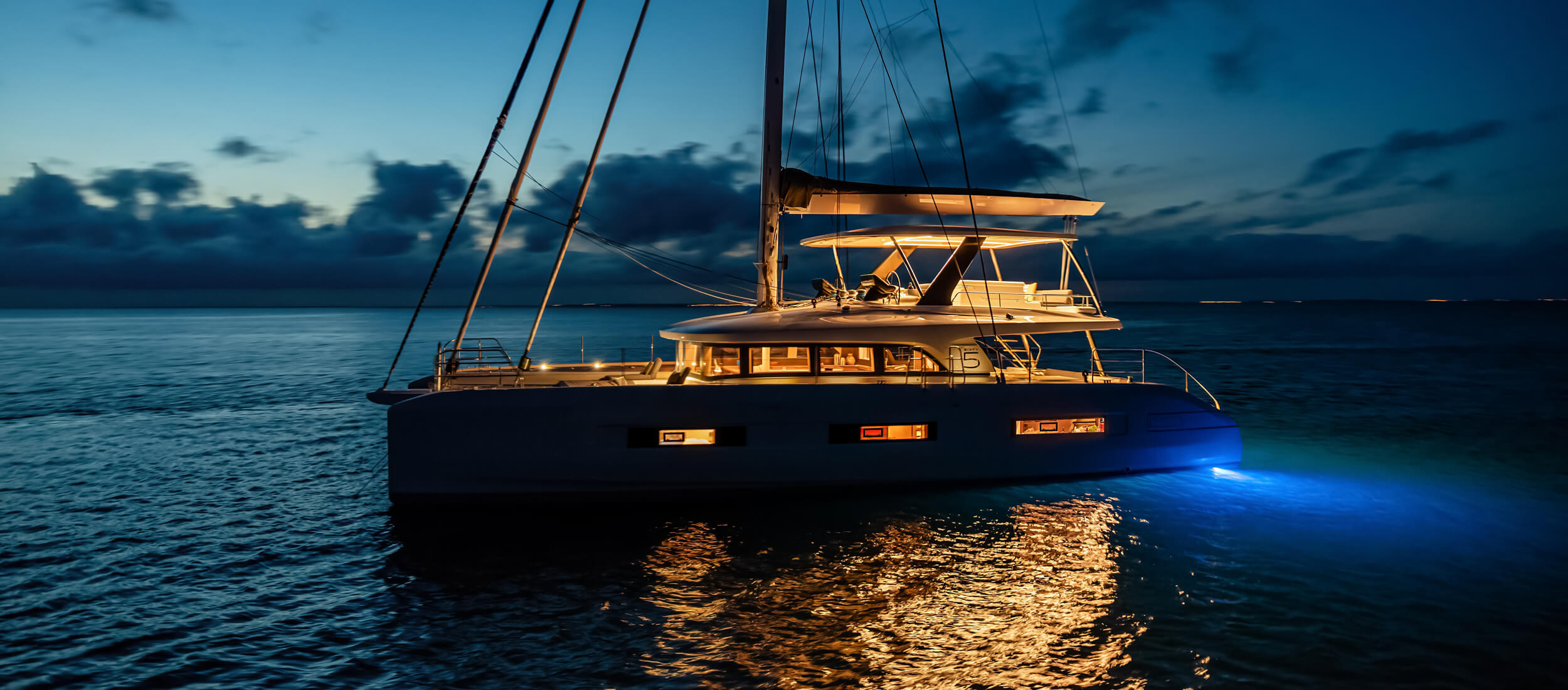 Athenian Yachts-A luxury catamaran anchored at dusk on calm water, beautifully illuminated with warm interior lights and glowing blue underwater lights beneath the hull.-Lagoon Catamarans for Sale